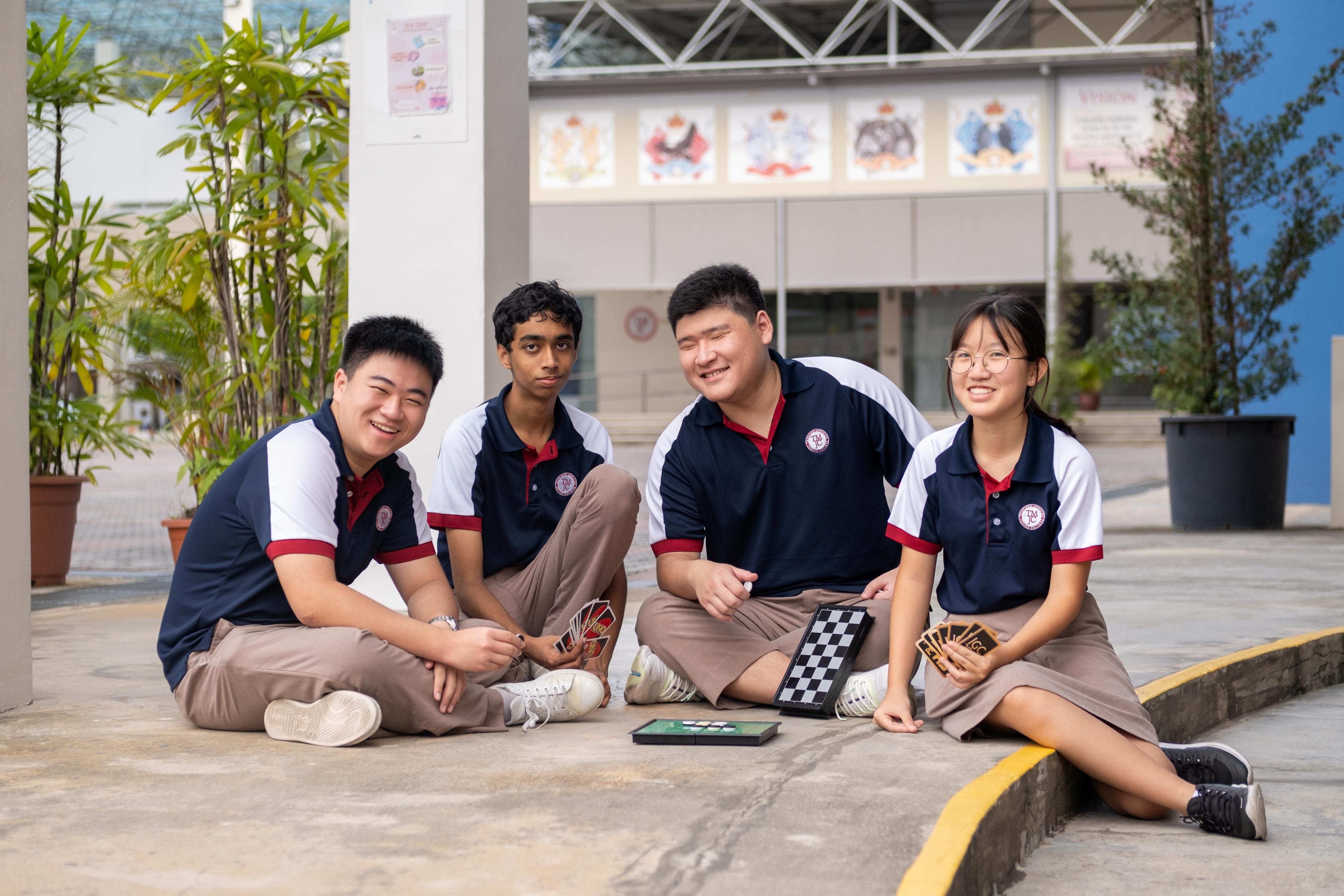 Four students in navy-and-white TMJC polo shirts sit casually on the ground outdoors in a school courtyard. Between them are a small chessboard and a hand of playing cards. They are smiling and relaxed, with potted plants and school building banners visible in the background.